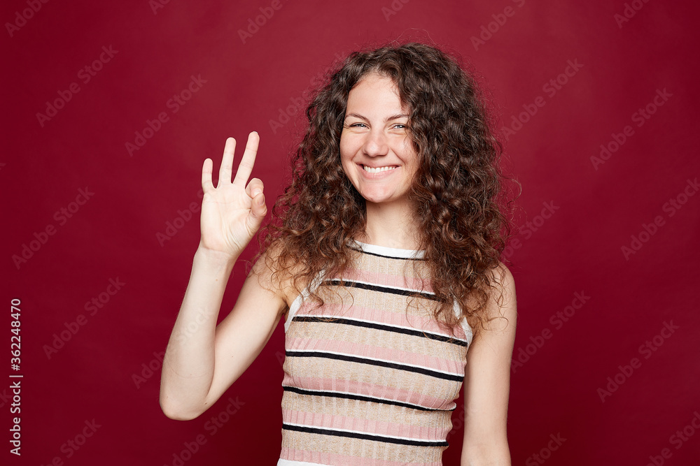 Attractive cheerful positive blue eyed female with toothy smile shows ok sing at camera, feeling good and confident. Pretty lCaucasian lady in striped shirt posing on crimson studio wall background. Attractive cheerful positive blue eyed female with toothy smile shows ok sing at camera, feeling good and confident. Pretty lCaucasian lady in striped shirt posing on crimson studio wall background.
