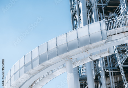 A close-up view of a contemporary construction of an industrial oil refinery or a modern fuel factory facility with a round bridge in front, multiple pipes, iron beams, and stairs, blue sky behind photo