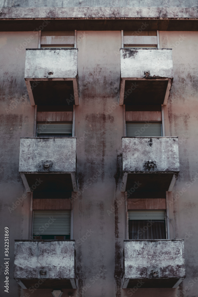 Vertical shot with a facade of a residential building: six balconies with single windows on each, some of them with partly closed blinds, a grungy wall with a blackened plaster, Lisbon, Portugal