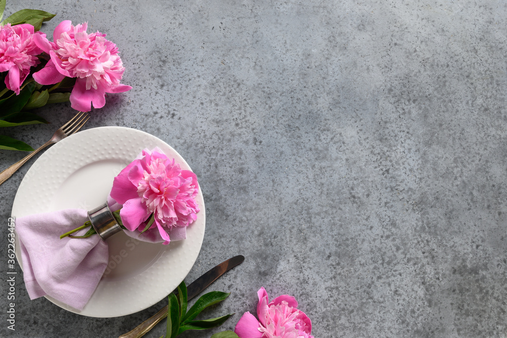 Festive table setting with pink peony flowers, white dishware, silverware on gray. Top view. Space for text.