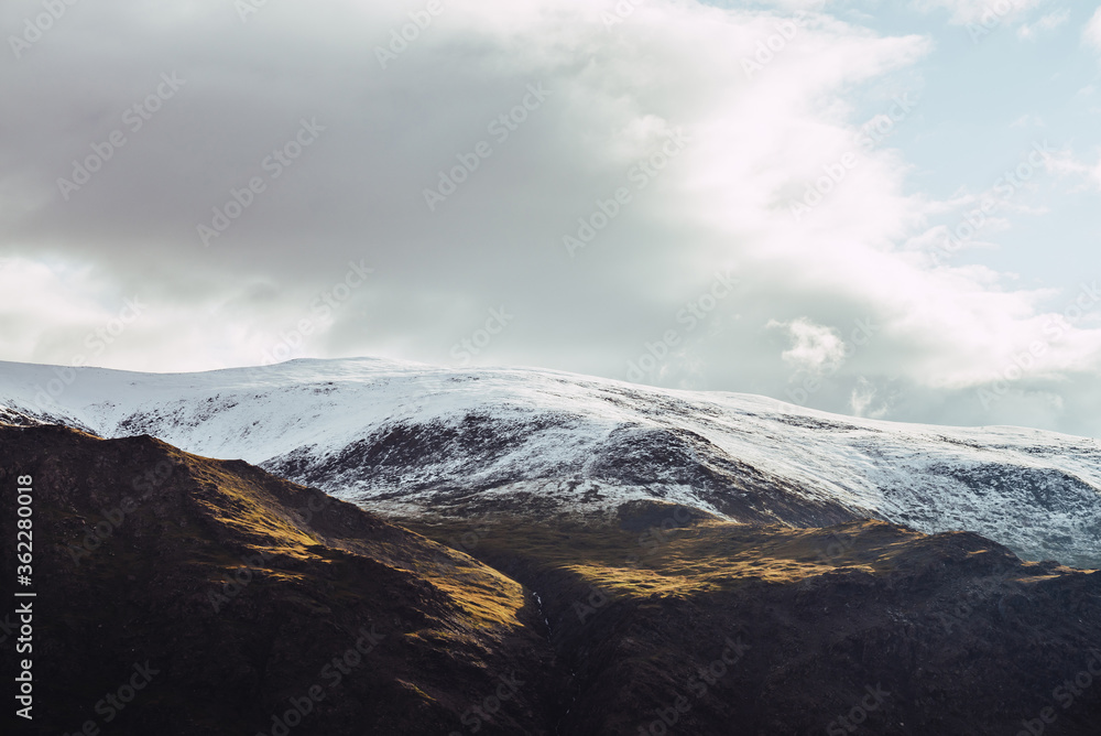 Atmospheric view to snowy mountains in sunlight under cloudy sky. Low clouds among big rocks with snow. Sunshine on white snow on mountain top. Scenic nature landscape with sunlight and low clouds.