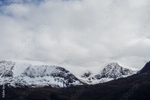 Great black mountains with white snow on tops and glaciers. Dramatic landscape with snowy mountains under cloudy gray sky. Atmospheric alpine scenery with snow on rocky mountains in overcast weather.