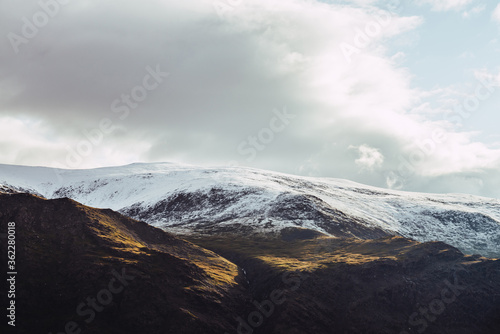 Atmospheric view to snowy mountains in sunlight under cloudy sky. Low clouds among big rocks with snow. Sunshine on white snow on mountain top. Scenic nature landscape with sunlight and low clouds.