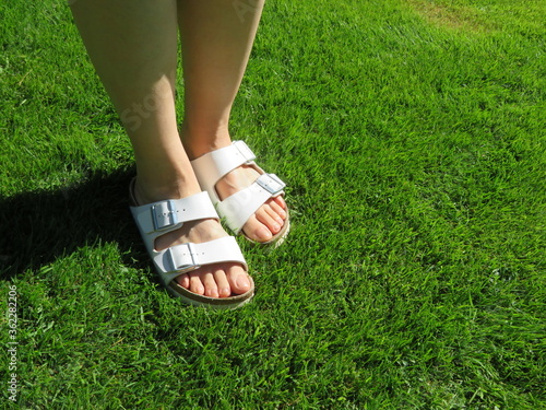 Closeup photo of woman feet in white sandals walking on grass during summer