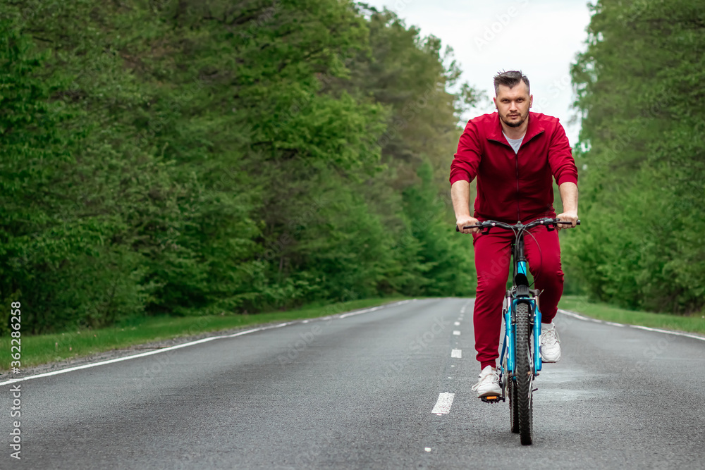 Fototapeta premium A man in a tracksuit on a bicycle rides on a road in the forest. The concept of a healthy lifestyle, cardio training. Copyspace.