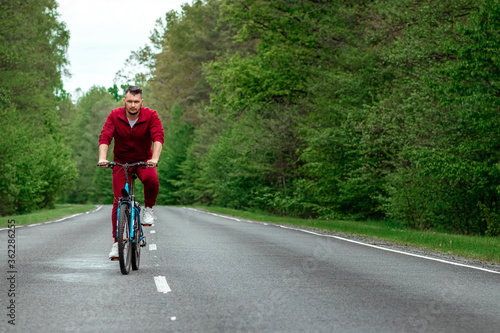 Fototapeta Naklejka Na Ścianę i Meble -  A man in a tracksuit on a bicycle rides on a road in the forest. The concept of a healthy lifestyle, cardio training. Copyspace.
