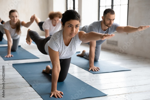 Concentrated young female indian trainer showing bird dog pose at group yoga class. Fit barefoot diverse people practicing body balancing exercise in Parsva Balasana position on mat indoors in gym.