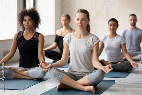 Smiling sporty professional european appearance female trainer leading yoga class to mixed race young people in studio club, sitting together in lotus position with folded in mudra gesture fingers.