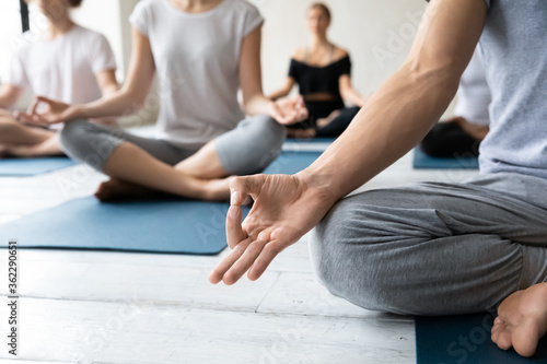 Close up young mindful guy puts folded in mudra gesture hand on knee, meditating or doing breathing exercises before start group class or finishing intensive workout with students in yoga studio.