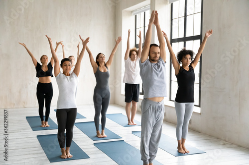 Front view group of focused happy young mixed race people in sportswear repeating surya namaskar exercises after professional male trainer barefoot on floor mat in modern yoga studio interior.