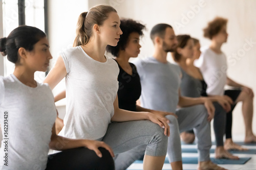 Group of young fit people standing in row in One Legged King Pigeon Pose II, stretching entire front body in Eka Pada Rajakapotasana II position together at group yoga class in modern sport club.