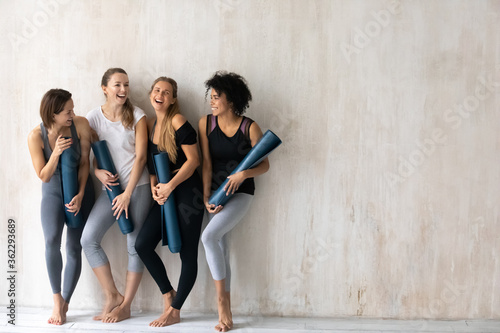 Full length emotional pretty young multiracial fit girls in activewear and floor mats in hands having fun before yoga class workout start, standing near wall with copy space for advertisement text.