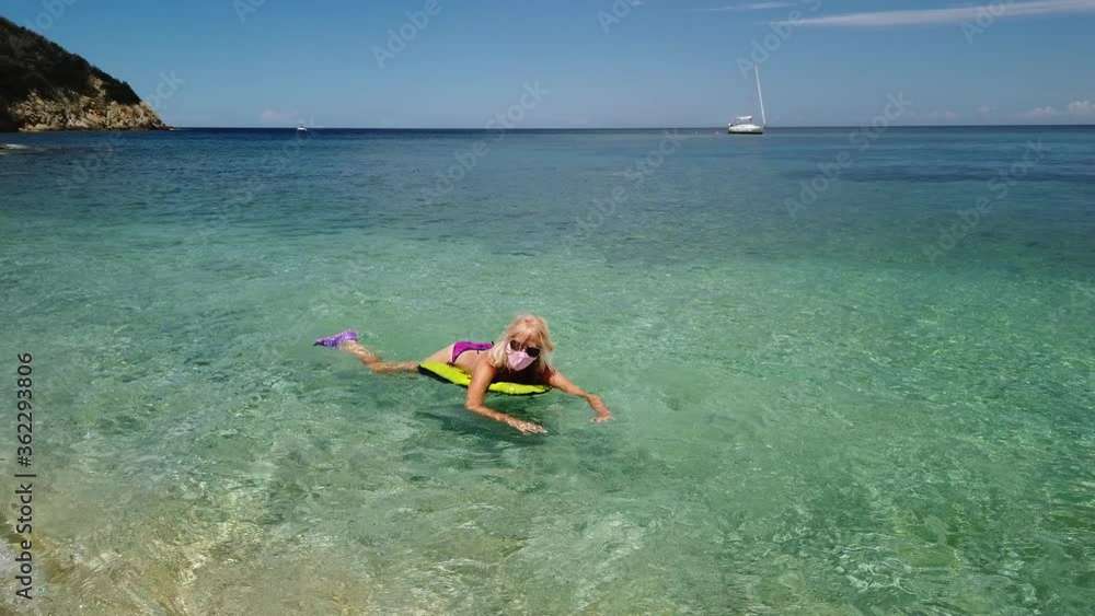 Woman swimming in Prunini beach on air mattress with surgical mask during Covid-19. Blonde woman having a bath in Elba island. Summer travel with Covid-19 pandemic in Italy. Woman swimming in Prunini beach on air mattress with surgical mask during Covid-19. Blonde woman having a bath in Elba island. Summer travel with Covid-19 pandemic in Italy.