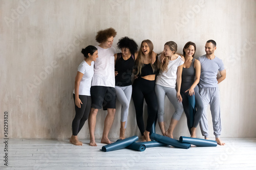 Full length happy young friendly mixed race diverse people in activewear cuddling standing barefoot near wall, feeling excited before yoga class start, relaxing after intensive workout finish indoors.