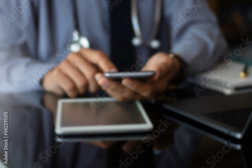 Blurred image of male doctor in blue shirt with tie, hand holding and using mobile smart phone with modern laptop computer, medical stethoscope and wireless mouse on the desk. Medical online concept.