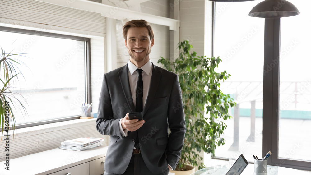 Portrait of smiling Caucasian businessman stand in own office using modern smartphone gadget, happy motivated young male CEO or boss holding cellphone gadget, posing at workplace, leadership concept Portrait of smiling Caucasian businessman stand in own office using modern smartphone gadget, happy motivated young male CEO or boss holding cellphone gadget, posing at workplace, leadership concept