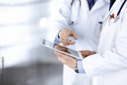 Two unknown doctors with stethoscopes discussing medical exam resoults, standing at hospital office. Physicians use a computer tablet for filling up medication names records. Perfect medical service