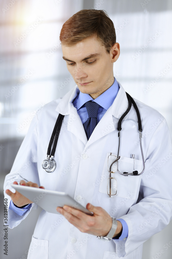Young doctor man is standing in a sunny hospital office, holding a computer tablet in his hands. Perfect medical service in clinic. Happy future in medicine and healthcare Young doctor man is standing in a sunny hospital office, holding a computer tablet in his hands. Perfect medical service in clinic. Happy future in medicine and healthcare