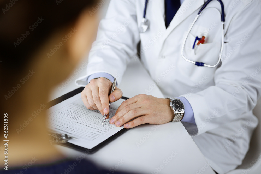 A doctor is talking to his patient, while sitting together at the desk in the cabinet in a hospital. Physician using clipboard for filling up medication history records. Perfect medical service in A doctor is talking to his patient, while sitting together at the desk in the cabinet in a hospital. Physician using clipboard for filling up medication history records. Perfect medical service in