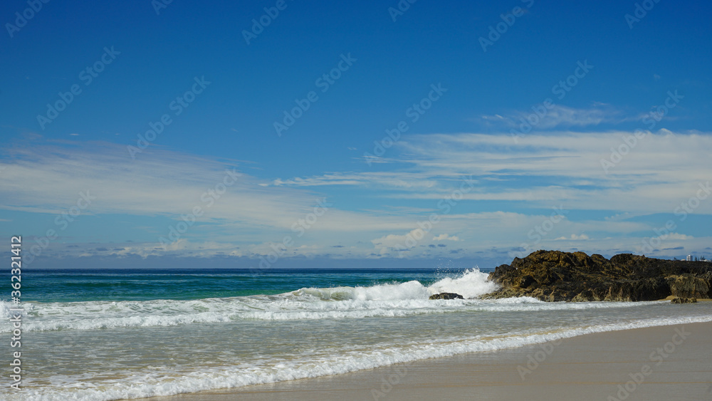 Seascape: surf breaking on rocks, with sandy beach in the foreground, clouds on the horizon, and blue sky above.