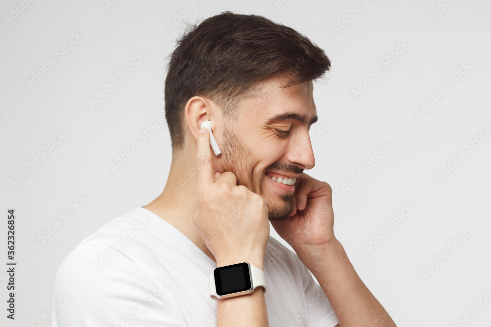 Side portrait of young handsome man in white t-shirt, wearing smartwatch and listening to music in wireless earphones, isolated on gray background Side portrait of young handsome man in white t-shirt, wearing smartwatch and listening to music in wireless earphones, isolated on gray background