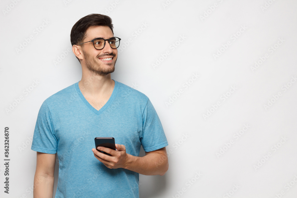 Banner of young aspired man standing in blue t-shirt holding phone in hand and looking away with dreamful smile, isolated on gray background