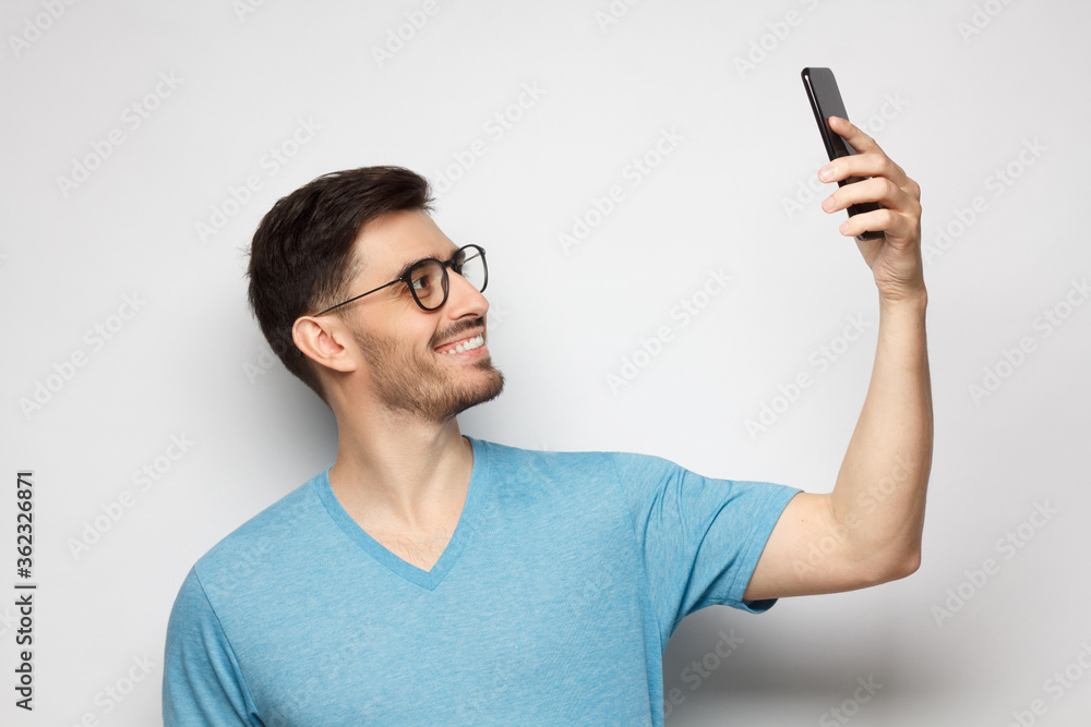 Young handsome man in blue tshirt holding phone and taking selfie photo, isolated on gray background Young handsome man in blue tshirt holding phone and taking selfie photo, isolated on gray background