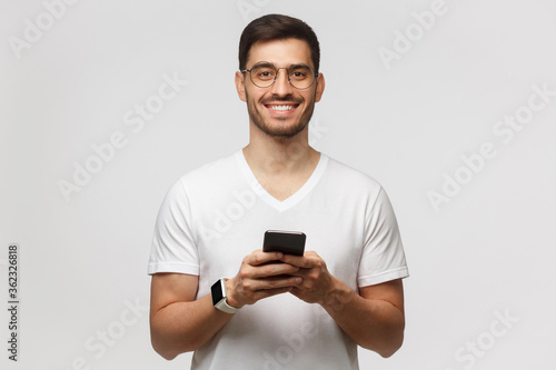Young handsome man in white t-shirt, smartwatch and round glasses, standing with phone in hands, isolated on gray background © Damir Khabirov