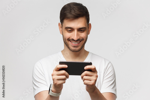 Young man holding phone in horizontal way as if watching video or playing video game, isolated on gray background
