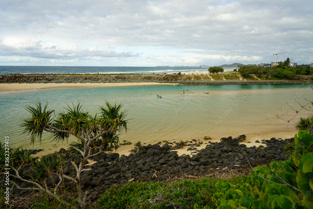 Looking across the creek and sea wall to the beach, with pandanus palm and rocky shore in the foreground and people kayaking, snorkelling and walking. Tallebudgera, Gold Coast, Queensland, Australia.