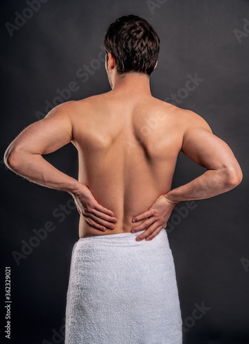 Handsome young bearded man isolated. Portrait of topless muscular man is standing on gray background. Man holding his back. Experiencing backache.