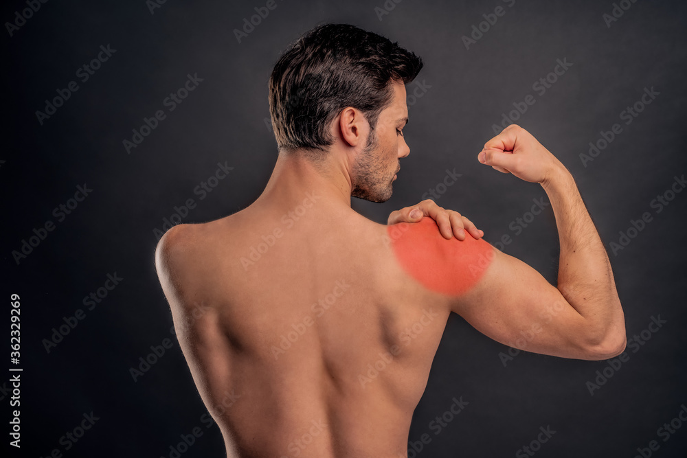 Handsome young bearded man isolated. Portrait of naked muscular man is standing on gray background. Back view of man holding his shoulder. Experiencing shoulder pain.