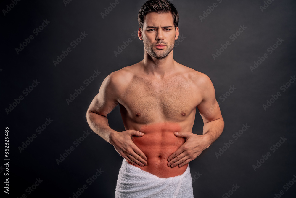 Fototapeta premium Handsome young bearded man isolated. Cropped image of shirtless muscular man is standing on gray background. Man holding his stomach. Experiencing stomach pain.