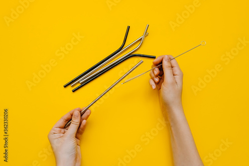 Human hand holding metallic straw, showing how to clean it with a special brush, on clean yellow background.