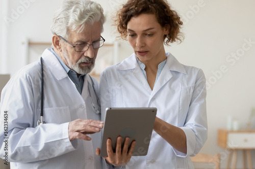 Concentrated man and woman doctors in white medical uniforms look at tablet screen discuss patient anamnesis or checkup results together, diverse GP talk brainstorm make decision using pad gadget