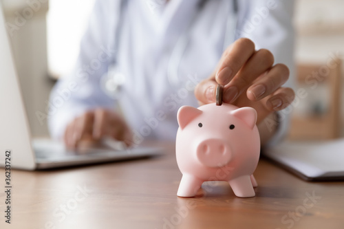 Crop close up of woman doctor hand put coin in piggybank, make monetary donation to clinic fund, female drop money contribution or volunteer charity in piggy bank for patient or hospital need care