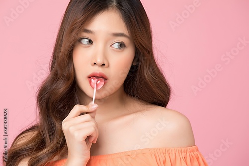 Young and cute Asian woman model holding a heart-shaped candy posing in front of pink studio background.