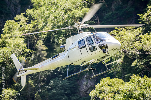 快晴の長野県松本市安曇の2300mの日本最大規模の氷河圏谷、北アルプス奥穂高岳直下の涸沢カールを飛行中の白い機体の一機のヘリコプター。背景は日光照りつける新緑の森。A helicopter at the cirque Karasawa on the east side of Mt.Okuhotakadake in the Northern alps.（東邦航空のヘリコプターJA6573を加工。）