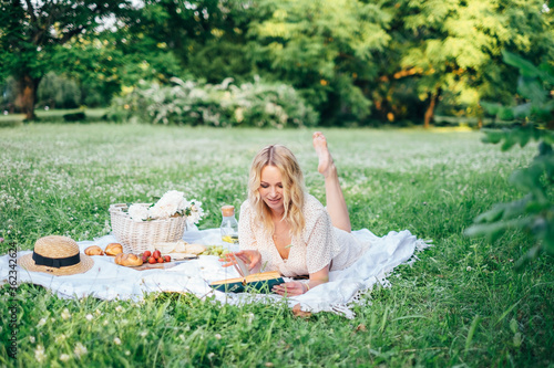 Young beautiful european blonde longhaired girl in white dress at nature.Spends the weekend outside the city, outdoors, a picnic in the park, relaxation, read book.Summer time Hold bascet with flowers © alex