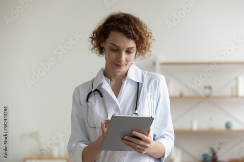 Young Caucasian female doctor or GP in white medical uniform use consult patient on tablet gadget, woman nurse or physician work on modern pad device, fill anamnesis, modern clinic hospital concept