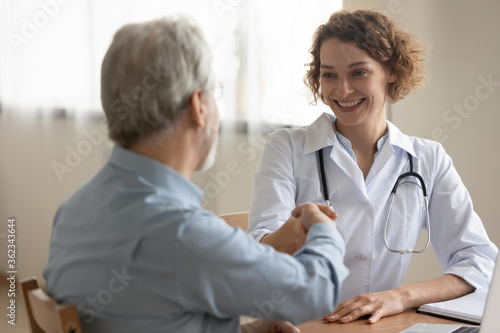 Smiling young female doctor or nurse shake hand get acquainted greeting with mature male patient in hospital, happy caring woman GP handshake senior man client at meeting, elderly healthcare concept