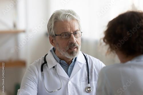 Old Caucasian male doctor in white medical uniform talk consult female patient at meeting in hospital, mature man GP or physician speak discuss illness or checkup with client at consultation in clinic
