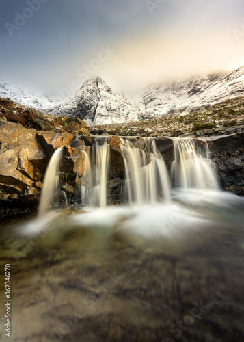 Beautiful Winter scene at the Fairy Pools on the Isle of Skye in the Scottish Highlands with snowcapped mountains and long exposure waterfall in the foreground.
