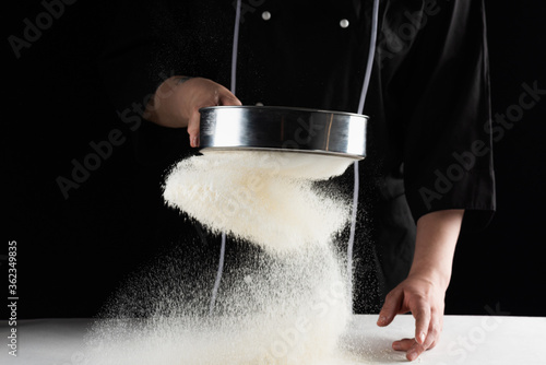 a dark background baker's hands sift white wheat flour over a sieve over photo