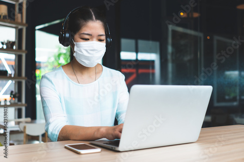 young asian woman wearing face mask and headphones and working remotely by using computer video call confernece at coworking space. social distancing and new normal lifestyle concept