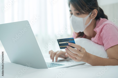 Sickness woman with face mask holding credit card and using computer laptop for online shopping on bed in morning at home. New normal, technology, ecommerce, digital banking and online payment concept