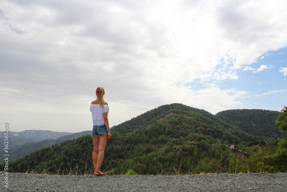 Naklejka premium A girl in shorts stands back and looks at the Troodos mountains from the observation platform of the Kikk monastery. Cyprus.