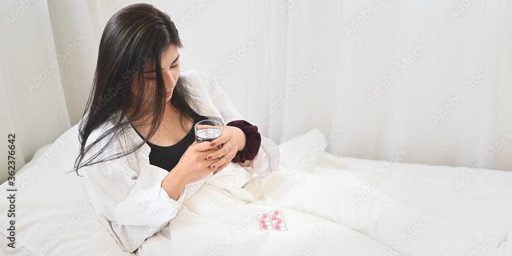 An ill woman is holding a glass of water after consuming a pill at the bed. An ill woman is holding a glass of water after consuming a pill at the bed.