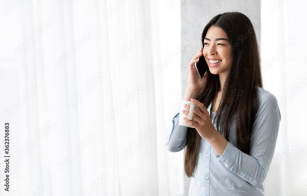 Pleasant call. Asian girl talking on cellphone and drinking coffee near window Pleasant call. Asian girl talking on cellphone and drinking coffee near window