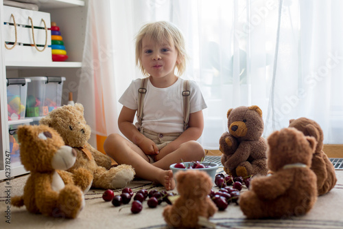 Toddler blond child, cute boy, eating cherries with teddy bears